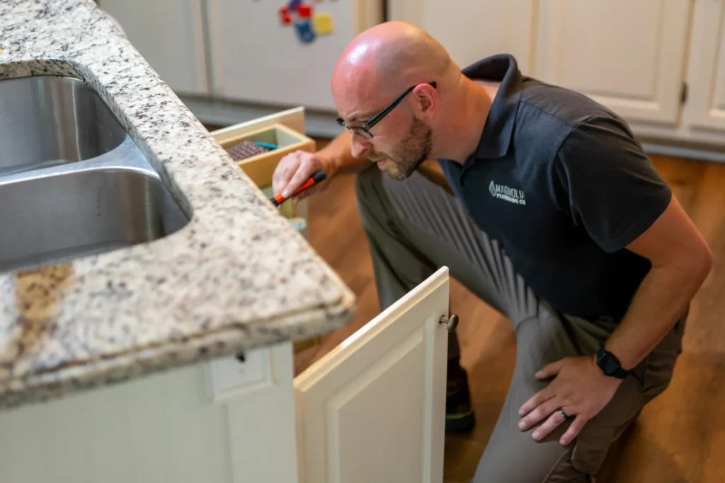 A man in glasses and a black polo shirt inspects the inside of a kitchen cabinet below a granite countertop, holding a flashlight and kneeling on the floor—a typical scene for Magnolia Plumbing's residential plumbing services in Memphis TN.