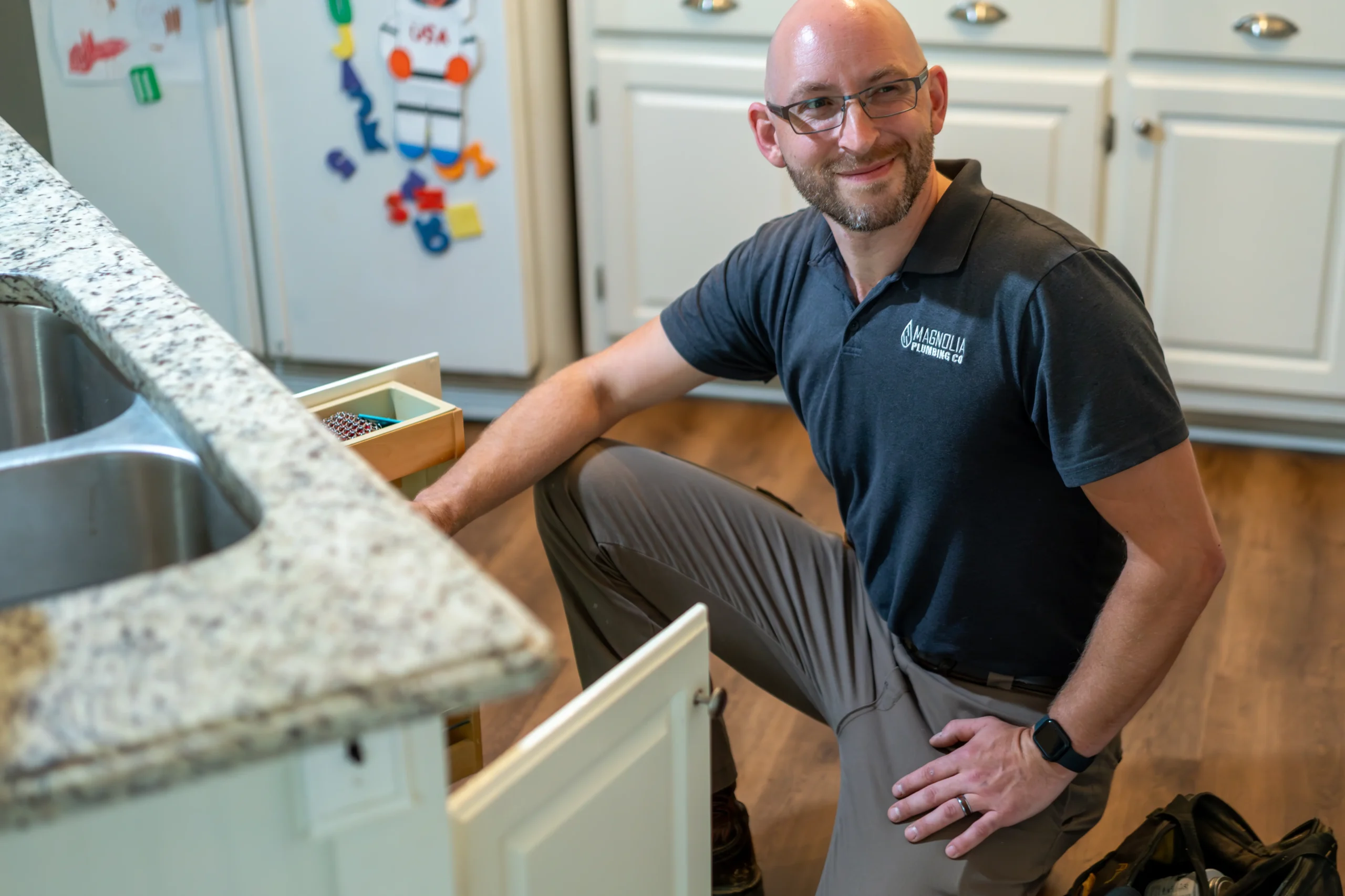 A man with glasses and a beard, wearing a dark polo shirt and khaki pants, kneels by an open kitchen cabinet, smiling while working on residential plumbing. The kitchen has white cabinets and a refrigerator with magnets—service by Magnolia Plumbing in Memphis TN.