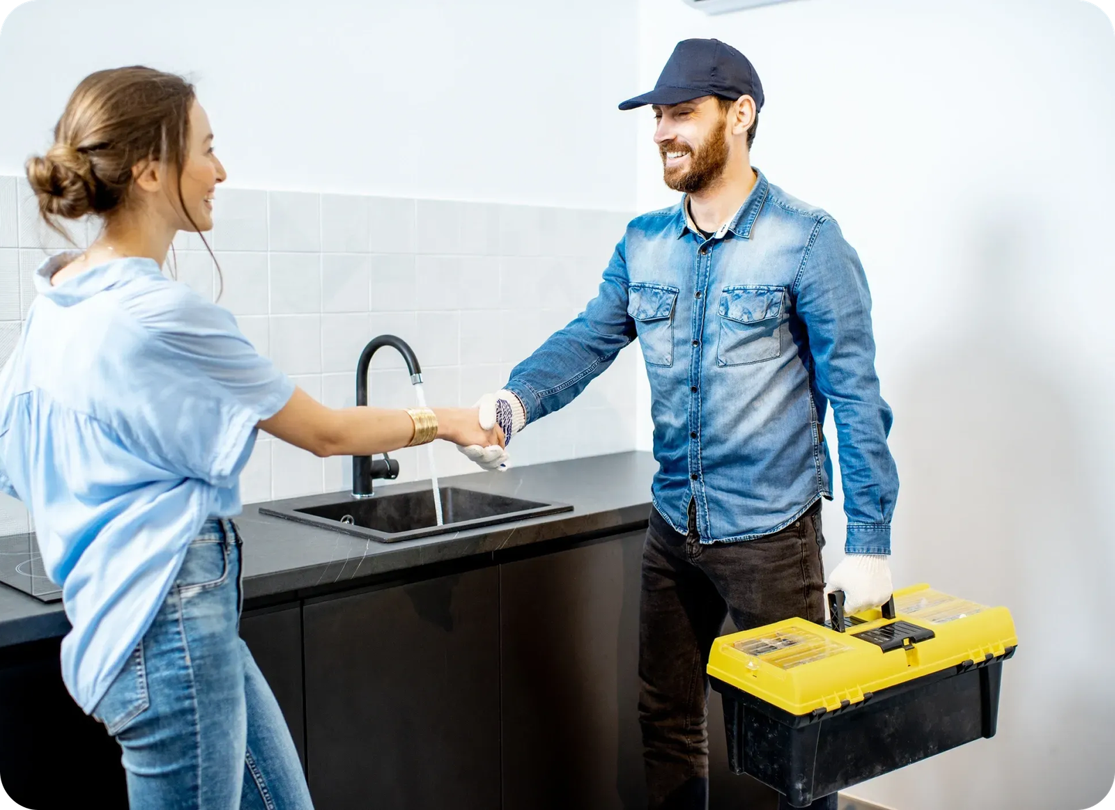 A smiling handyman from Magnolia Plumbing in a denim shirt and cap, holding a yellow toolbox, shakes hands with a woman in her Memphis TN kitchen near the sink—your trusted choice for residential plumbing solutions.