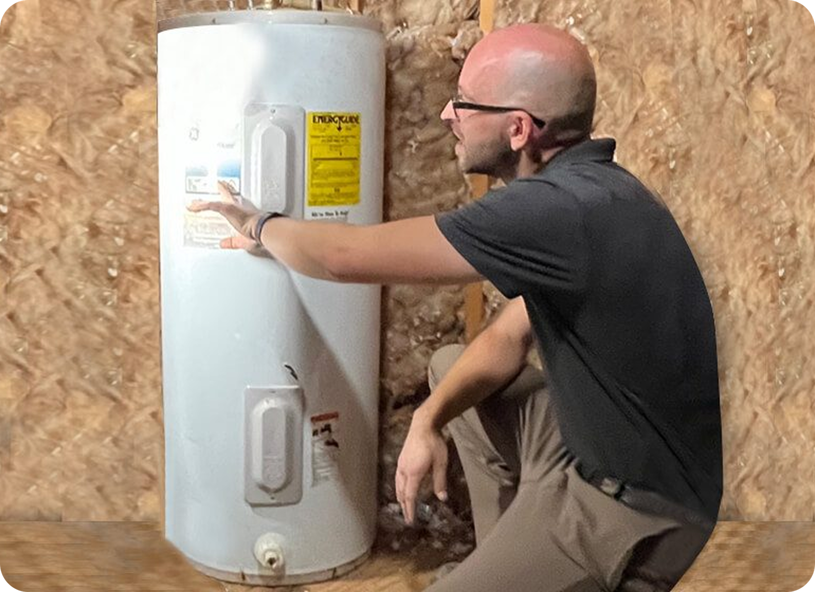 A man in glasses and a black shirt crouches next to a white water heater in an unfinished room, pointing at a label on the heater—showcasing expert residential plumbing in Memphis, TN, with insulation visible on the walls behind him.