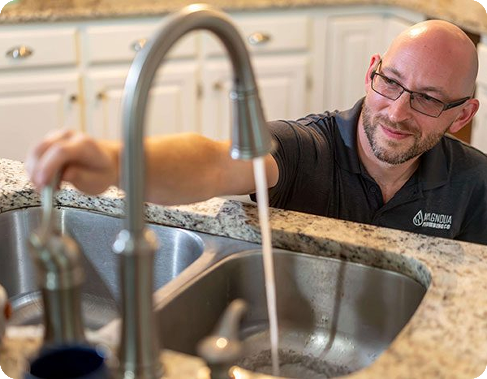 A man in glasses and a Magnolia Plumbing shirt demonstrates residential plumbing skills by turning on a kitchen faucet, letting water flow into a double sink set in a granite countertop. White cabinets complete the modern Memphis, TN kitchen scene.