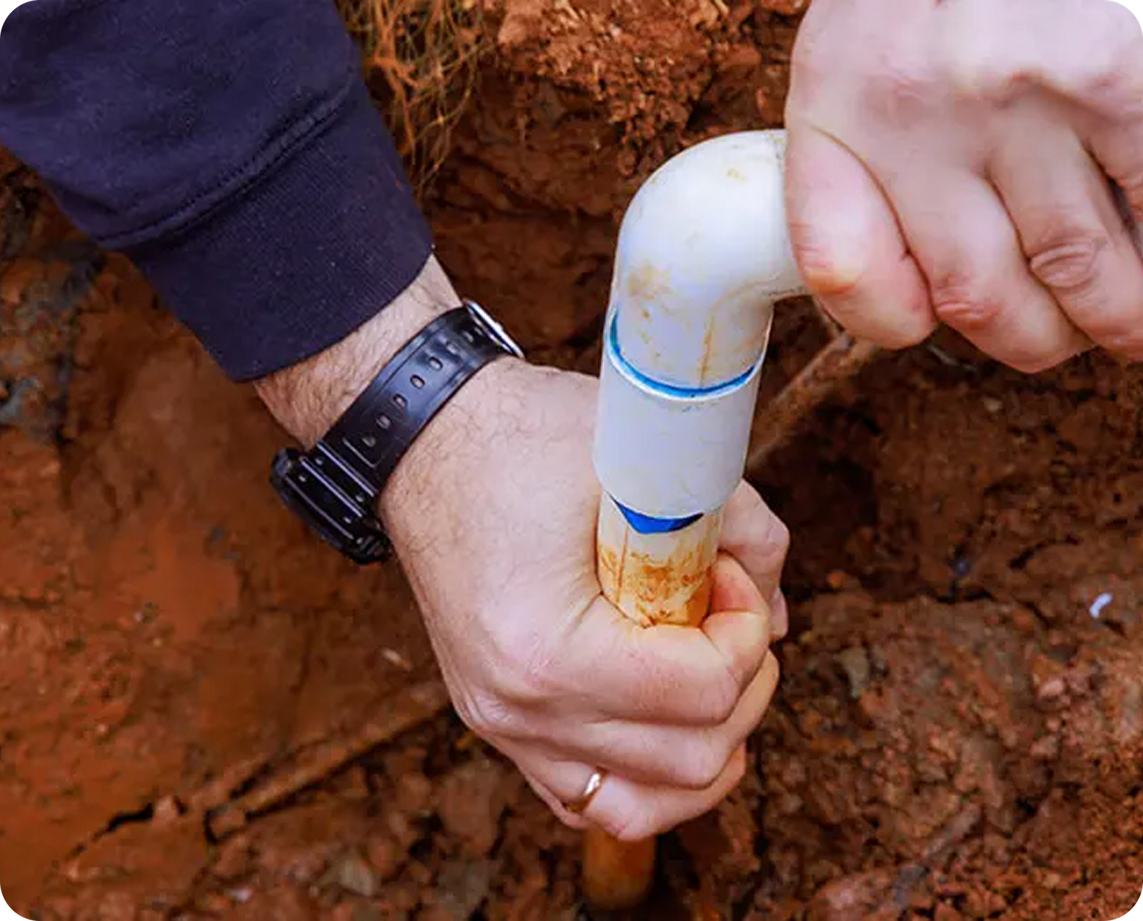 Close-up of a person’s hands tightening a white PVC pipe fitting outdoors in reddish soil, wearing a black watch and dark long-sleeve shirt—showcasing Magnolia Plumbing’s expert residential plumbing work in Memphis, TN.