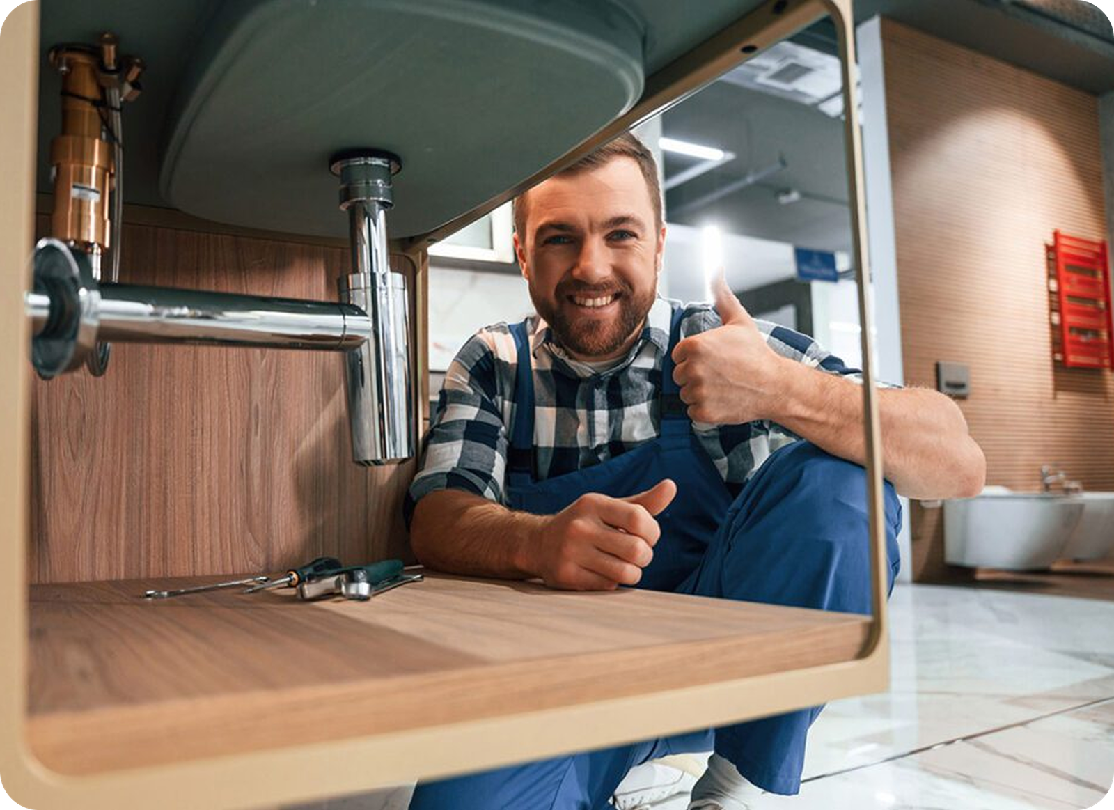 A plumber from Magnolia Plumbing in blue overalls smiles and gives a thumbs-up while crouching under a sink, with tools laid out nearby in a modern Memphis, TN bathroom—your trusted choice for residential plumbing solutions.