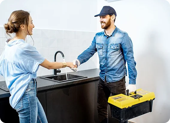 A woman shakes hands with a smiling Magnolia Plumbing repairman in a denim shirt and cap, who is holding a yellow tool box in a modern Memphis, TN kitchen near a sink.