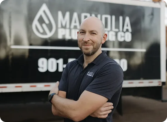 A bearded man in a black polo shirt and smartwatch stands with arms crossed in front of a Magnolia Plumbing Co. truck, showcasing the company logo and phone number—your trusted choice for residential plumbing in Memphis, TN.