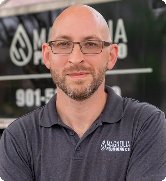 A man with glasses and a shaved head is wearing a gray Magnolia Plumbing Co. polo shirt, standing in front of a company vehicle featuring the Magnolia Plumbing logo and phone number, representing residential plumbing services in Memphis TN.